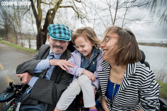 A white woman with short hair, wearing glasses and a black-and-white striped jacket, and a white man wearing a blue and black checkered newsboy cap, both wheelchair users, are smiling and laughing. A white toddler sits between them, perched on the armrests of their wheelchairs. They are outdoors near a lake, with trees and a road visible in the background.