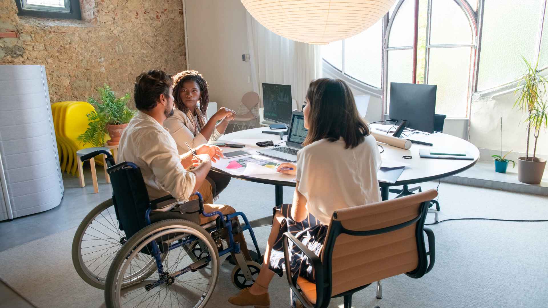 Three people are having a meeting in a modern office space. One person is in a wheelchair, another is seated on an office chair, and the third is leaning forward while speaking. Laptops, papers, and phones are on the round table. The room has large arched windows, potted plants, and exposed brick walls.